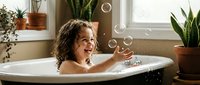 Happy mixed-race toddler girl with curly hair playing with soap bubbles in white bathtub surrounded by green houseplants in bright bathroom interior.