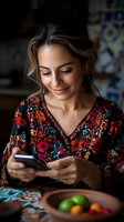 Young Hispanic woman in floral embroidered dress smiling while using smartphone at home. Natural lighting creates intimate atmosphere with fruit bowl in foreground.