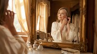 Middle-aged Caucasian woman in white robe applying skincare cream while looking in ornate vintage mirror during morning beauty routine in luxury bathroom.