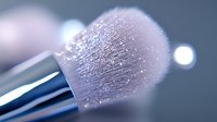 Professional makeup brush with soft pink bristles covered in shimmering powder particles against blurred background for beauty and cosmetics applications.