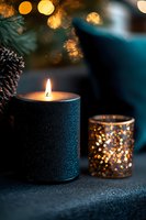 Glowing black candle and decorative glass holder with sparkling bokeh lights and pine cone create cozy winter atmosphere against dark background.