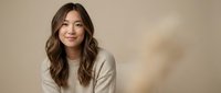 Young Asian woman with long wavy brown hair smiling warmly in beige sweater against neutral background for lifestyle and portrait photography.