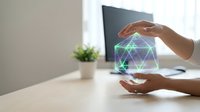 Hands protecting glowing geometric network hologram above laptop in modern office workspace representing cybersecurity and data protection technology.