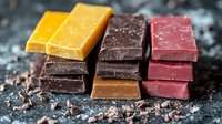 Stacked pieces of artisanal chocolate bars in different colors and flavors on dark background with cocoa crumbs, macro food photography for confectionery marketing.