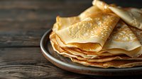 Freshly made thin crepes stacked on rustic ceramic plate against dark wooden background, golden brown pancakes with delicate texture and folds.