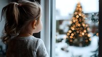 Young girl in gray sweater looking through window at Christmas tree with bokeh lights while snow falls outside, creating nostalgic winter holiday atmosphere.