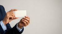 Male professional with medium brown skin tone wearing dark business suit holding blank white card or certificate in both hands against neutral background for corporate messaging.