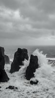 Dramatic black and white seascape featuring towering dark rock formations battered by powerful crashing waves and white sea spray under stormy skies. Suitable for nature, travel, and environmental design projects.