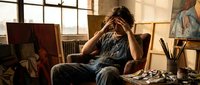 Young male artist with dark hair sitting in creative studio workspace, holding head in hands showing artistic frustration or contemplation surrounded by easels and paintings.
