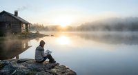 Person reading book by misty lake at sunrise with wooden cabin, peaceful morning meditation and solitude retreat for mental wellness and relaxation.
