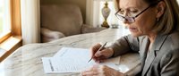 Senior businesswoman with glasses reviewing financial documents at home office desk near window for retirement planning and investment decisions.