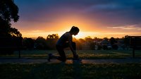 Athlete in athletic stance on grass field during golden hour sunset, silhouetted against vibrant orange and purple sky, ready position for sports training or outdoor fitness workout.