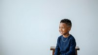 Young African American boy wearing navy blue long-sleeve shirt, seated on wooden chair against white background, smiling while looking to side, studio portrait for advertising and design projects.