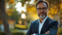 Confident mature Caucasian businessman with gray beard and glasses smiling outdoors in park during golden hour sunset, arms crossed, wearing navy suit.