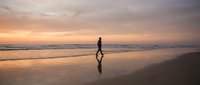 Silhouette of male surfer walking along sandy beach at sunset carrying surfboard, golden hour coastal scene for travel and lifestyle content.