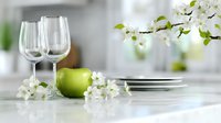 Elegant table setting with empty wine glasses, green apple and delicate white spring blossoms on light surface, creating fresh and minimalist dining atmosphere.