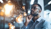 Young African American businessman in glasses contemplating innovative ideas, surrounded by glowing light bulbs representing creativity and inspiration in modern office space.
