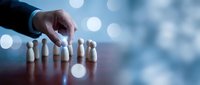 Businessman hand selecting wooden peg from row of figures on table with bokeh background, representing recruitment, leadership selection, and human resources management.
