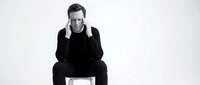 Young Caucasian businessman in black suit sitting on white cube with hands on face showing stress or contemplation on isolated white background.