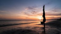 Silhouetted female athlete performing handstand on beach at sunset, practicing yoga or acrobatics by ocean waves during golden hour.
