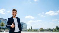 Young Caucasian male professional in dark blazer and white shirt giving thumbs up gesture outdoors with modern office buildings and blue sky background, representing success and approval.