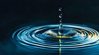 Water drop splash creating concentric ripples on dark blue surface with golden light reflections, captured in high speed macro photography showing liquid crown formation.