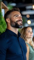 Handsome bearded man in navy shirt smiling and looking up with joy in modern restaurant setting with blurred woman and warm lighting atmosphere.