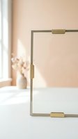 Minimalist glass photo frame with gold brass clips on white table, soft peach wall and dried flowers in background, modern home decor.