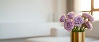 Lavender lisianthus blooms arranged in a gold vase on a table, soft sunlit interior background with copy space for text.