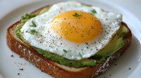 Sunny side up egg on avocado toast with fresh herbs and black pepper seasoning, served on rustic bread. Close up view of breakfast preparation.