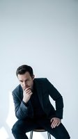 Thoughtful businessman in dark suit sitting on stool with hand on chin in contemplative pose against clean white background for professional use.