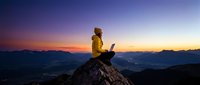 Person in yellow jacket sitting on mountain peak with laptop at sunset, working remotely with valley landscape below.