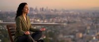 Young professional woman meditating on rooftop terrace overlooking city skyline at sunset. Business executive practicing mindfulness and wellness routine.
