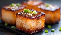 Caramelized pork belly cubes with crispy golden-brown top, tender meat, garnished with fresh chopped green onions served on dark plate. Perfect for food blogs.