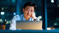 Young Asian businessman looking frustrated while working late at laptop in dark office with bokeh lights background, showing signs of stress and burnout.
