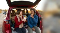 Young couple enjoying hot drinks from thermos during autumn road trip sitting in open car trunk at sunset creating cozy travel moments together.