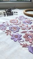 Floral embroidery with purple and pink flowers on white linen fabric, embroidery hoop and lavender sprigs in background, handmade needlework craft.