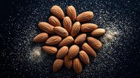Fresh whole almonds arranged in spiral pattern on dark background with scattered flour dust creating dramatic contrast and texture for food photography and packaging design.