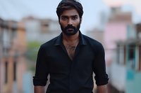 Young South Asian man with beard wearing black button-up shirt stands confidently against urban background in evening light, expressing serious contemplative mood.