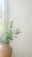 Lily of the valley and fern bouquet in a clay vase against a soft textured wall in warm natural light.