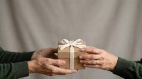 Hands exchanging wrapped gift box with cream ribbon bow against neutral background, suitable for holiday, celebration, giving, and generosity concepts.