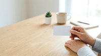 Woman holding blank white business card at wooden desk with succulent plant, coffee mug, and notebook for corporate branding and marketing purposes.