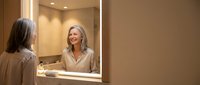 Mature woman with gray hair smiling at reflection in bathroom mirror during morning skincare routine in modern home interior with warm lighting.