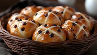 Fresh hot cross buns with raisins and white icing in rustic woven basket, traditional Easter holiday baking, golden brown glazed surface, close up view.