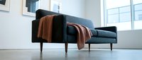 Modern dark blue upholstered sofa with wooden armrests and mid-century legs, styled with rust-colored throw blanket in minimalist bright interior space.