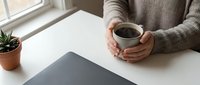 Woman in cozy sweater holding coffee mug at white desk with laptop and plant, perfect for remote work and home office lifestyle concepts.