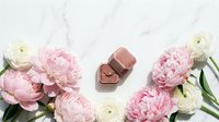 Elegant engagement ring in pink velvet box surrounded by peonies and ranunculus on marble, romantic wedding flat lay.