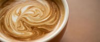 Creamy coffee latte with artistic foam swirl pattern in white ceramic cup on wooden surface, perfect for cafe menu and beverage marketing materials.