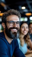 Happy bearded man with glasses smiling in modern office environment with colleagues in background for business and workplace concepts.