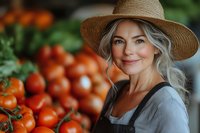 Mature Caucasian woman with gray hair and straw hat smiling at farmers market, wearing apron near fresh red tomatoes display, natural organic produce seller.